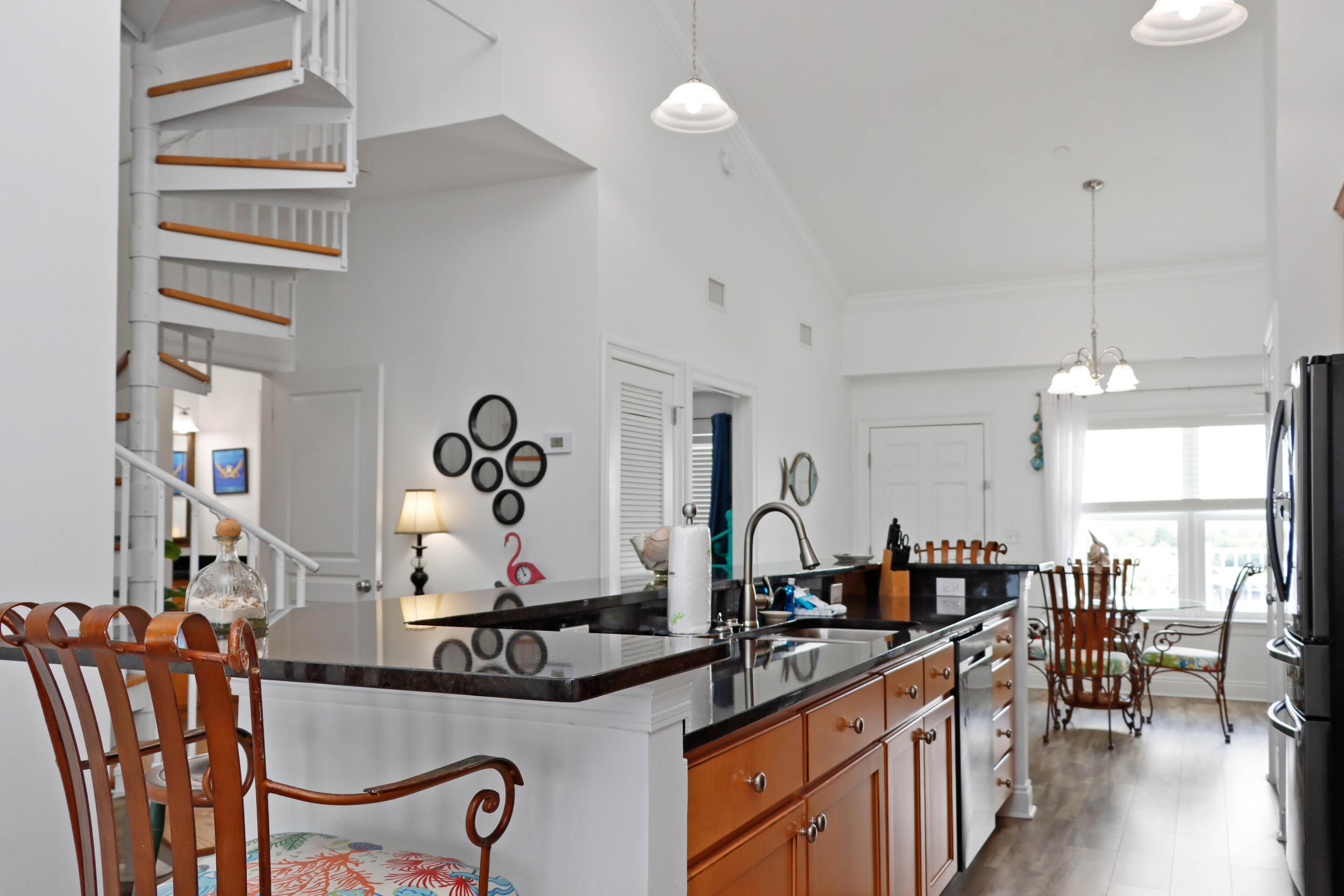 A spacious kitchen with a central island, wooden chairs, and a staircase leading to an upper level.