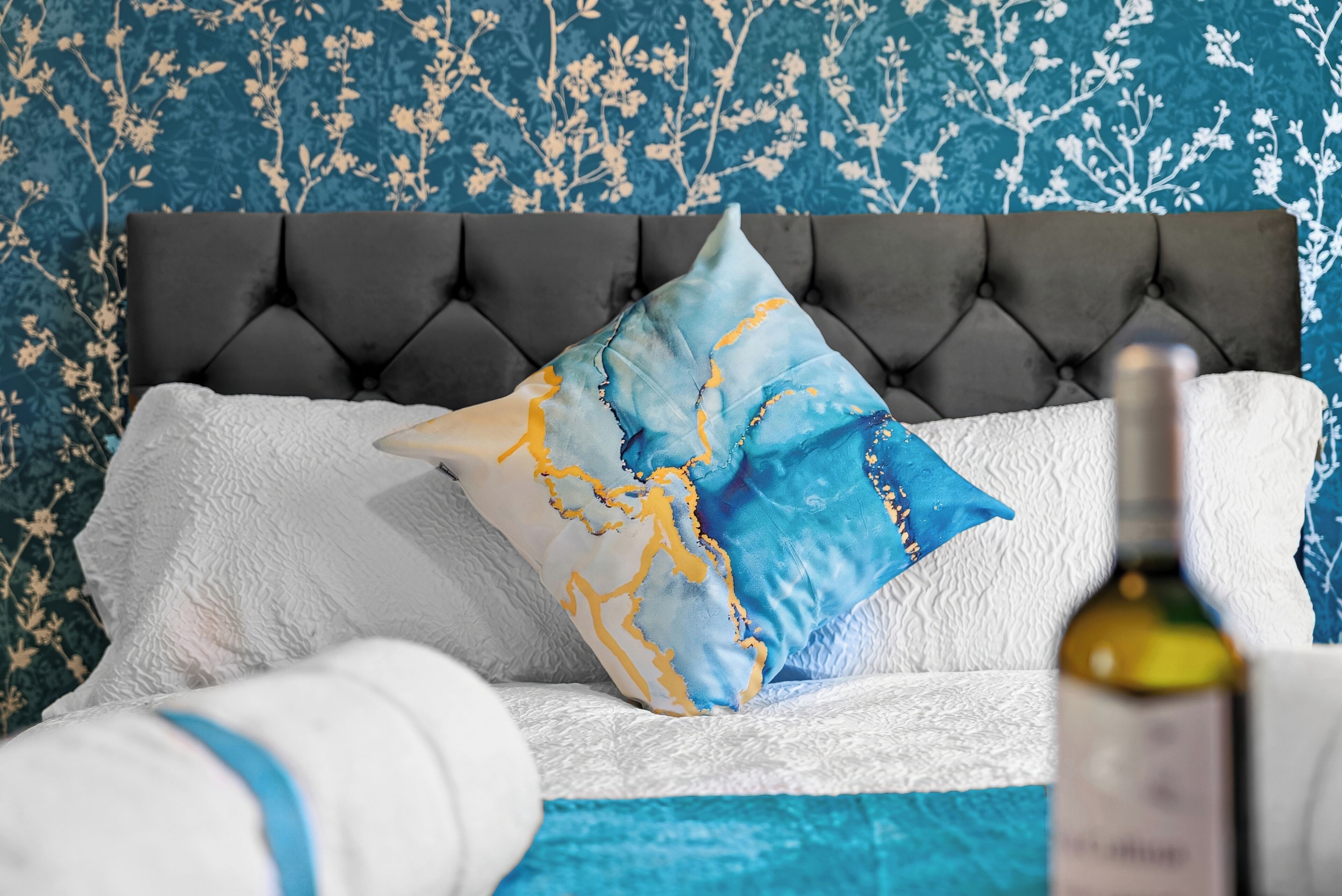 A bed with a tufted headboard, white bedding, and decorative pillows. A bottle of wine is visible on the bedside table.
