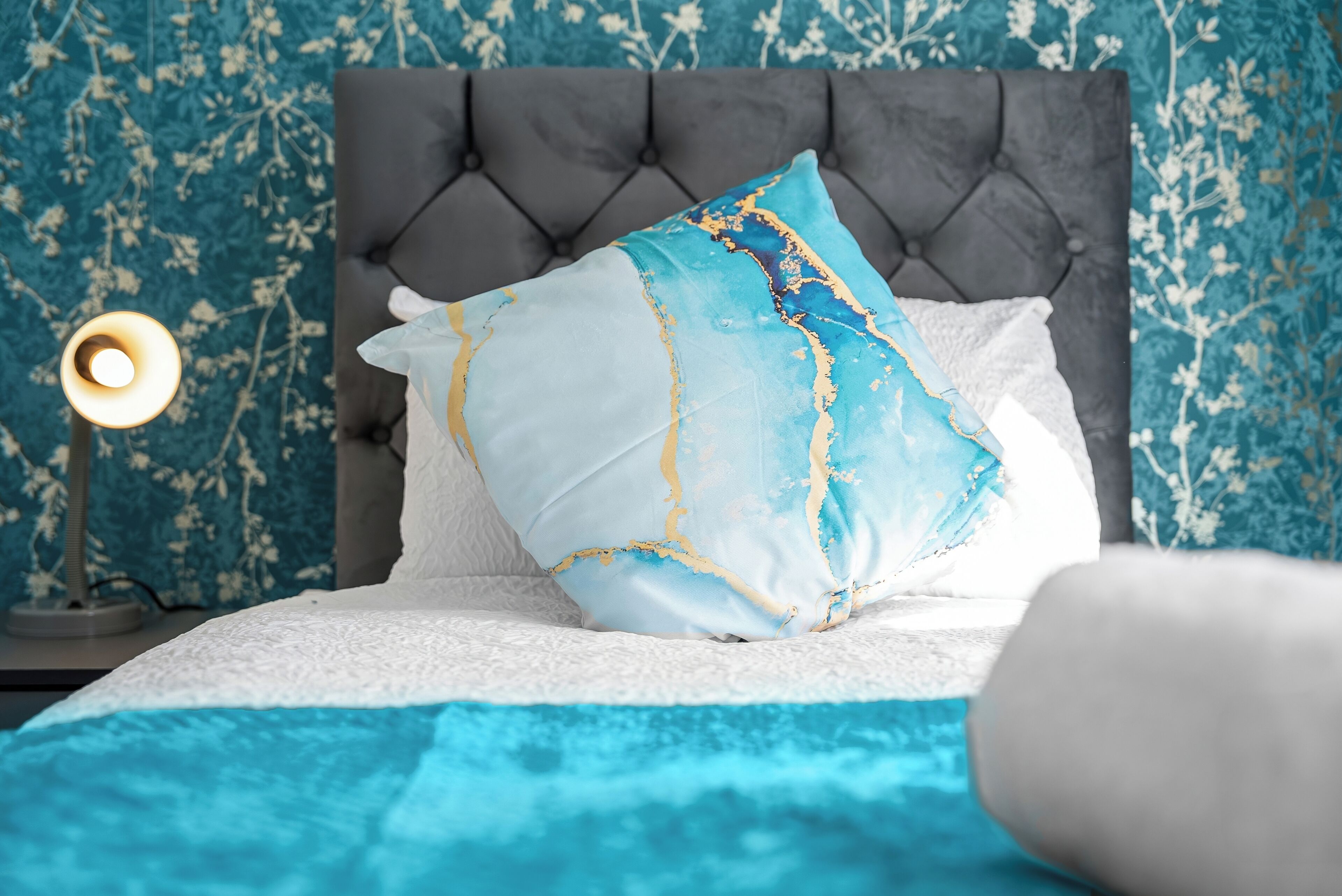 A bed with a tufted headboard, a blue and white patterned wall, and a bedside lamp.