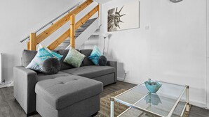 A modern living room with a grey sectional sofa, a glass coffee table, a staircase with wooden railings, and a wall clock. - Cozy 3 bedroom house near hospital (Coventry)