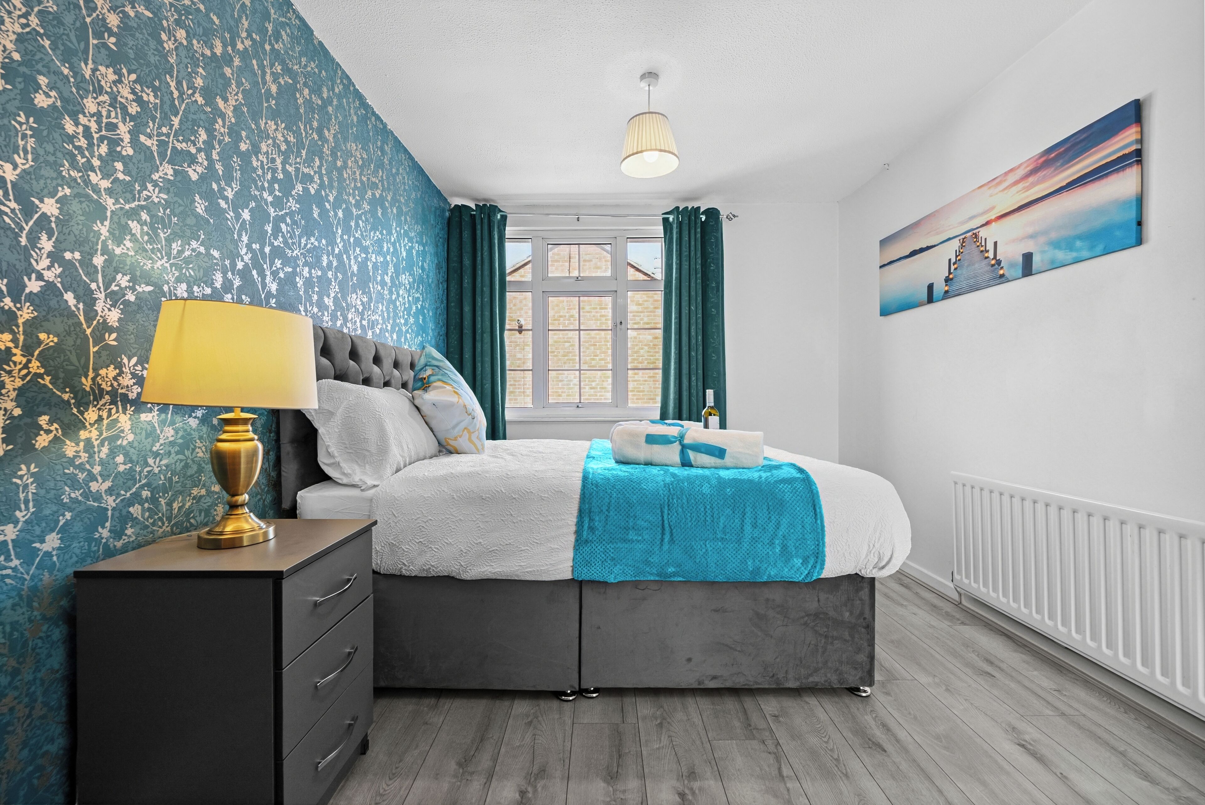 Une chambre à coucher comprenant un lit, une table de chevet, une lampe, une fenêtre avec des rideaux, un radiateur et un tableau mural.