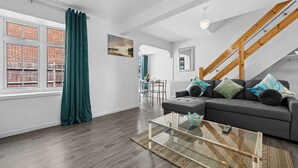 A modern living room with a grey sofa, a glass coffee table, and a view of a brick building through large windows. - Cozy 3 bedroom house near hospital (Coventry)