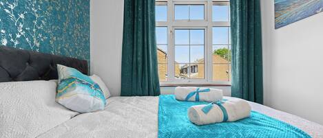 A bedroom with a large bed, teal curtains, a view of buildings outside, and a painting on the wall.