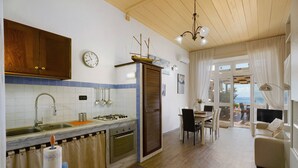Una cocina con estufa, pileta y muebles, un comedor con sillas y una mesa, y vista al exterior a través de un vidrio.