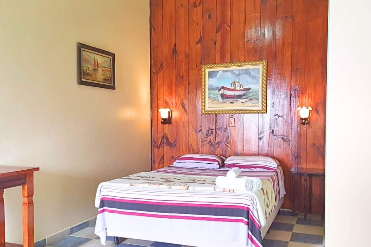 A bedroom with a wooden wall, a bed with striped bedding, two wall-mounted lamps, and a framed picture of a boat.