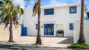 A white building with blue doors and windows, surrounded by palm trees and a paved area.