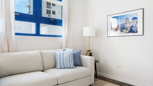 Un salon avec un canapé beige, un coussin à rayures bleues et blanches, une lampe blanche et un tableau encadré représentant un paysage côtier.