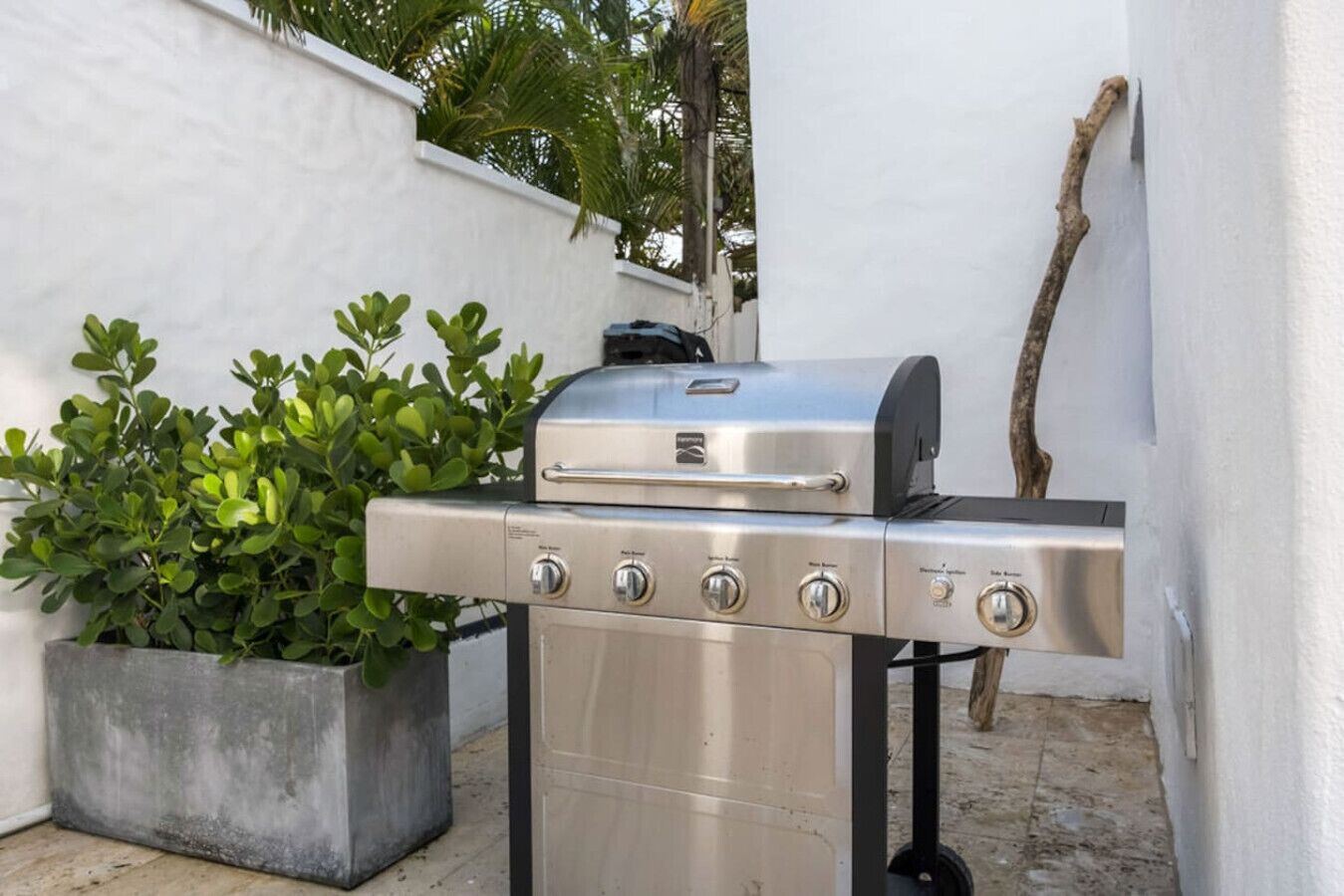 Un barbecue en acier inoxydable doté de quatre brûleurs et d’une étagère latérale, installé à l’extérieur contre un mur blanc, avec une plante en pot à proximité.