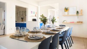A dining area with a long table set for six, surrounded by chairs, in a modern interior with white walls and a kitchen in the background.