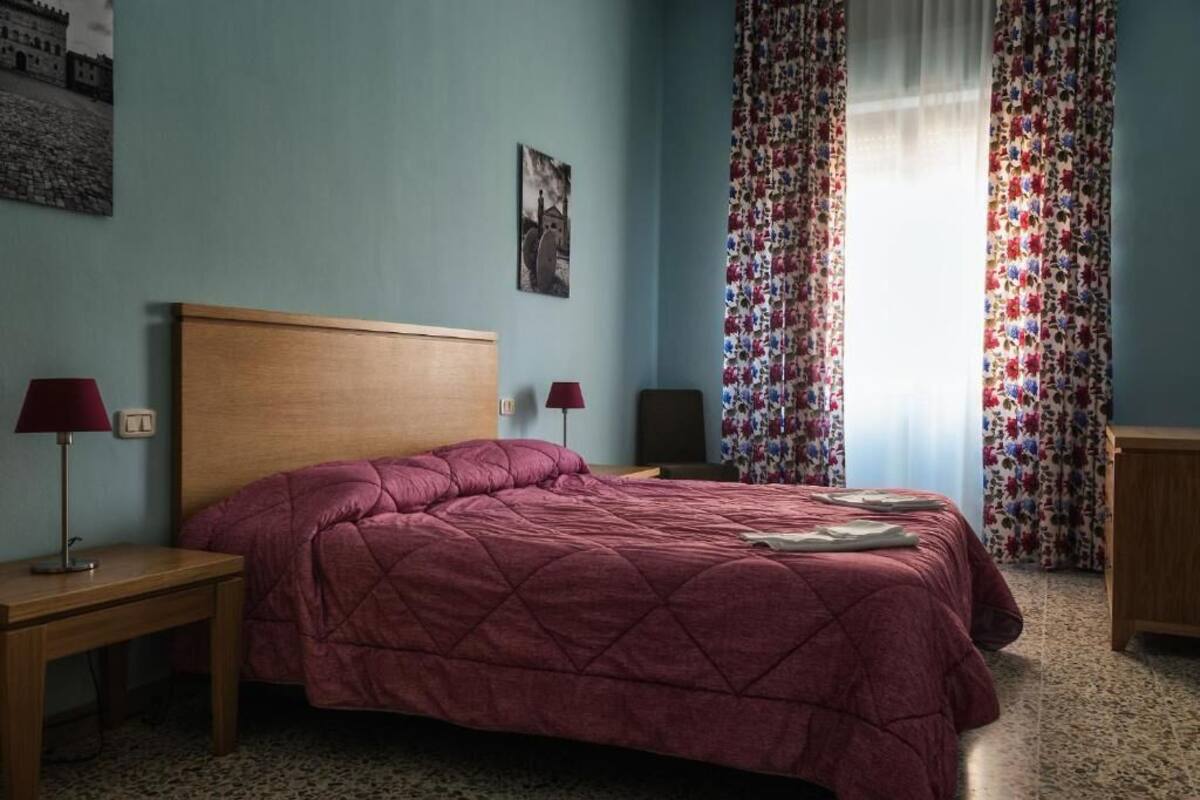 A bedroom with a wooden headboard, a red quilted bedspread, two bedside lamps, and a floral curtain.