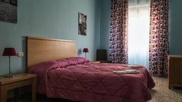 A bedroom with a wooden headboard, a red quilted bedspread, two bedside lamps, and a floral curtain.