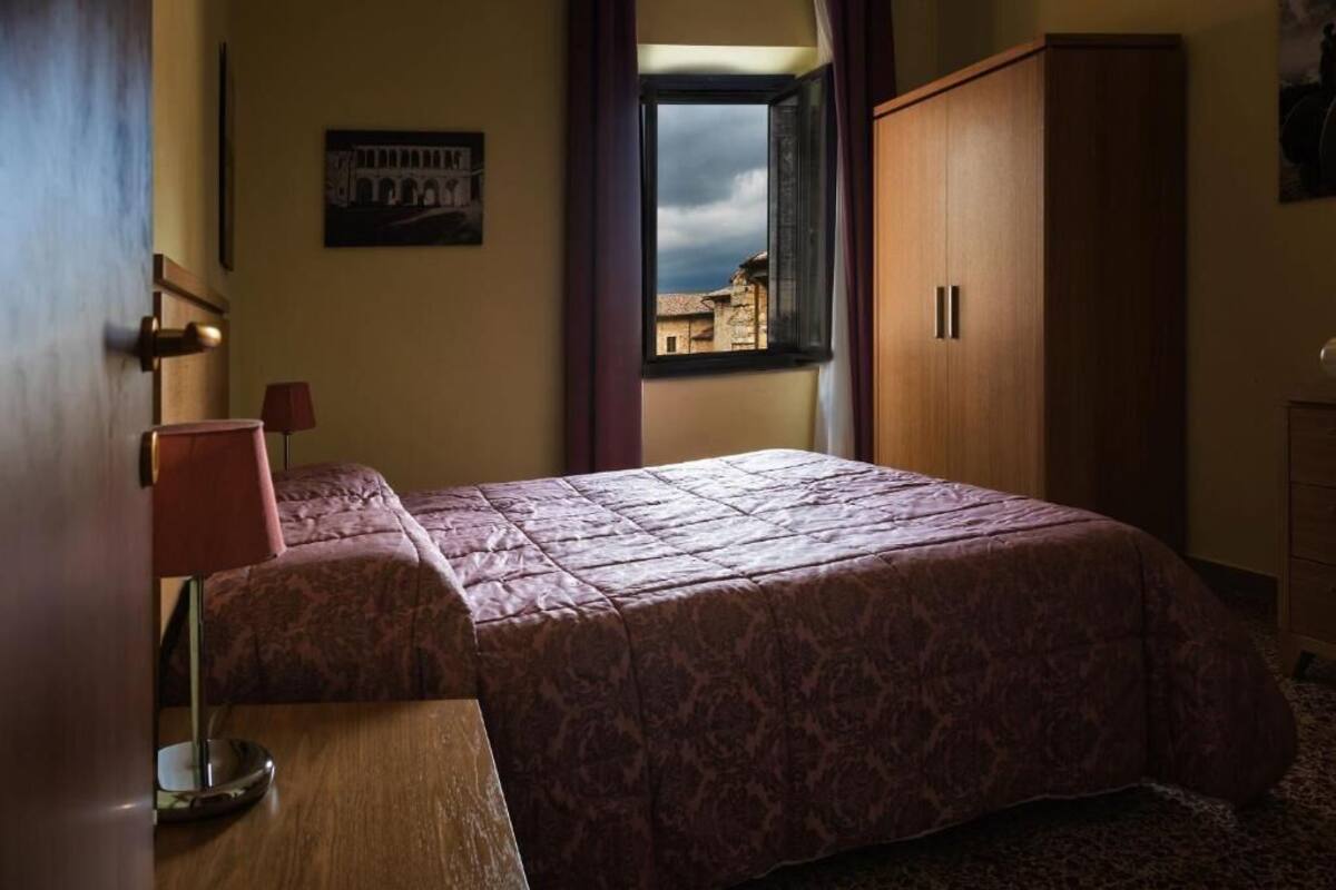 A bedroom with a bed, a wooden wardrobe, a window with a view of buildings, and a lamp.