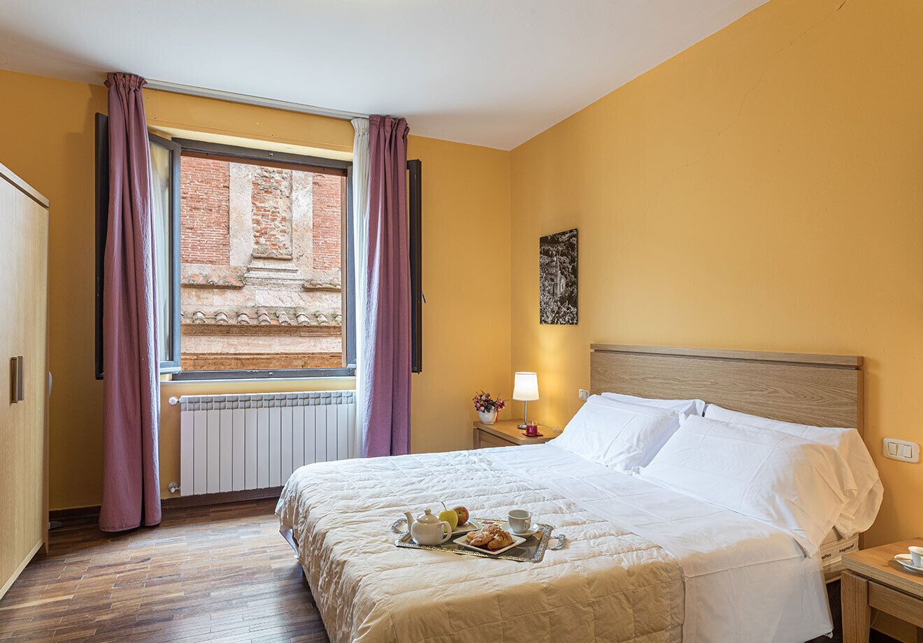 Ein Hotelzimmer mit Bett, einem fenster mit Vorhängen, einem Heizkörper, einem Nachttisch mit Lampe und einem Wandbild.