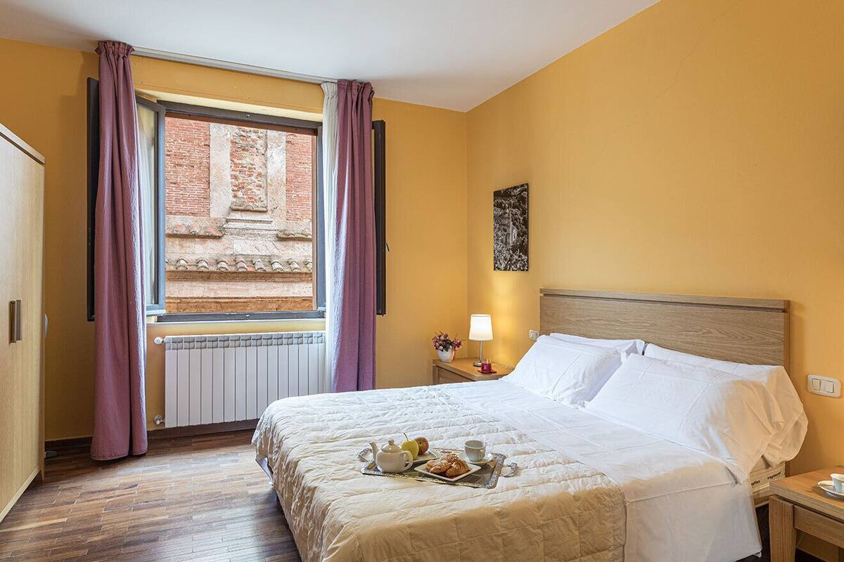 A hotel room with a bed, a window with curtains, a radiator, a nightstand with a lamp, and a wall-mounted picture.