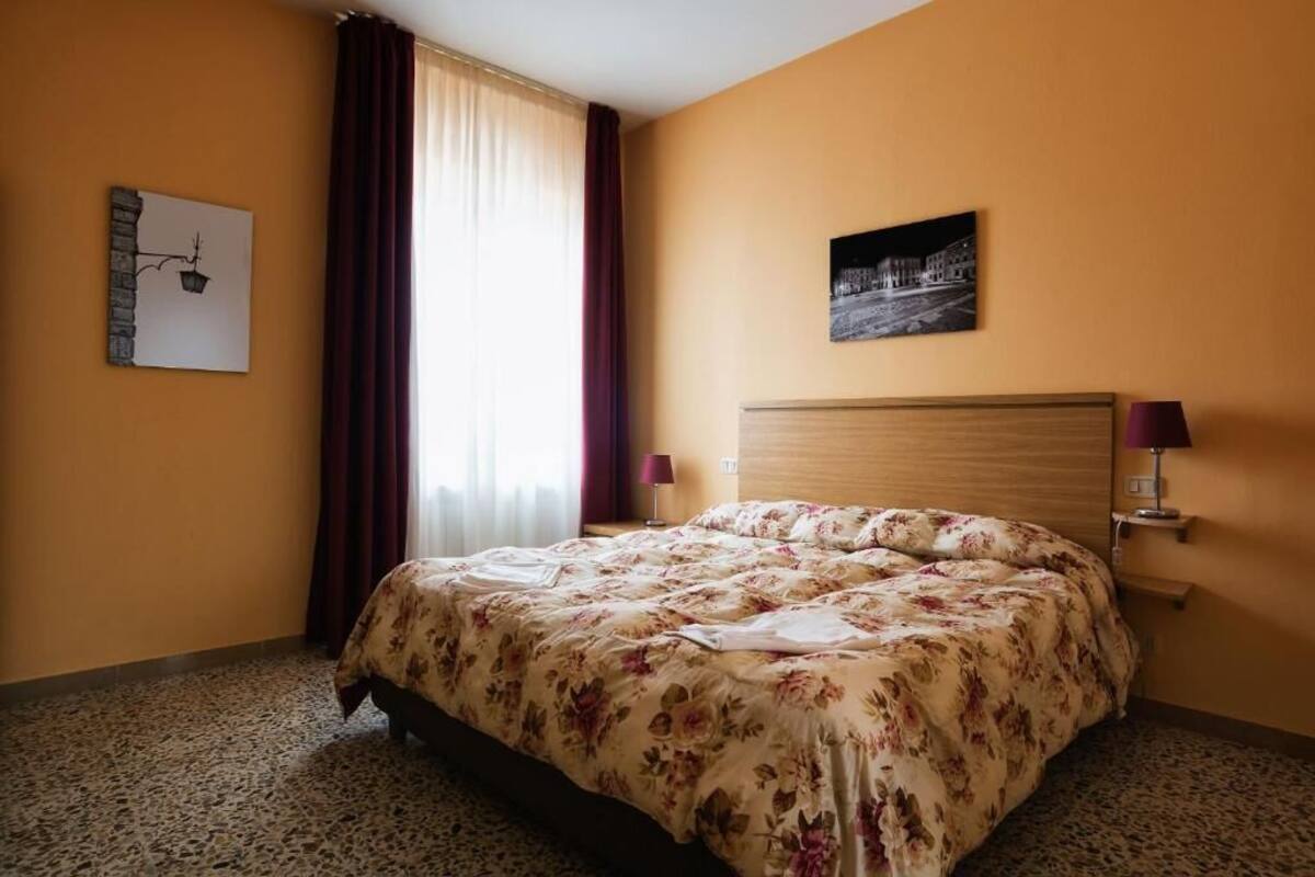 A hotel room with a bed, a nightstand, a lamp, a window with curtains, and a wall art piece.
