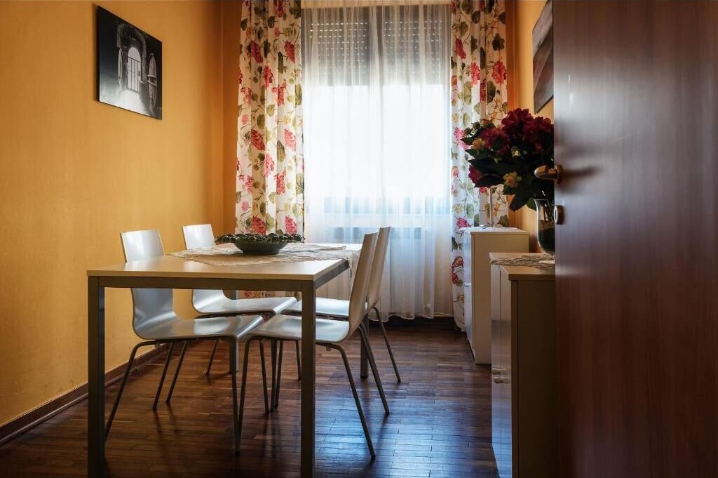 Uma área de refeições com uma mesa e cadeiras, uma janela com cortinas florais e um vaso com flores.