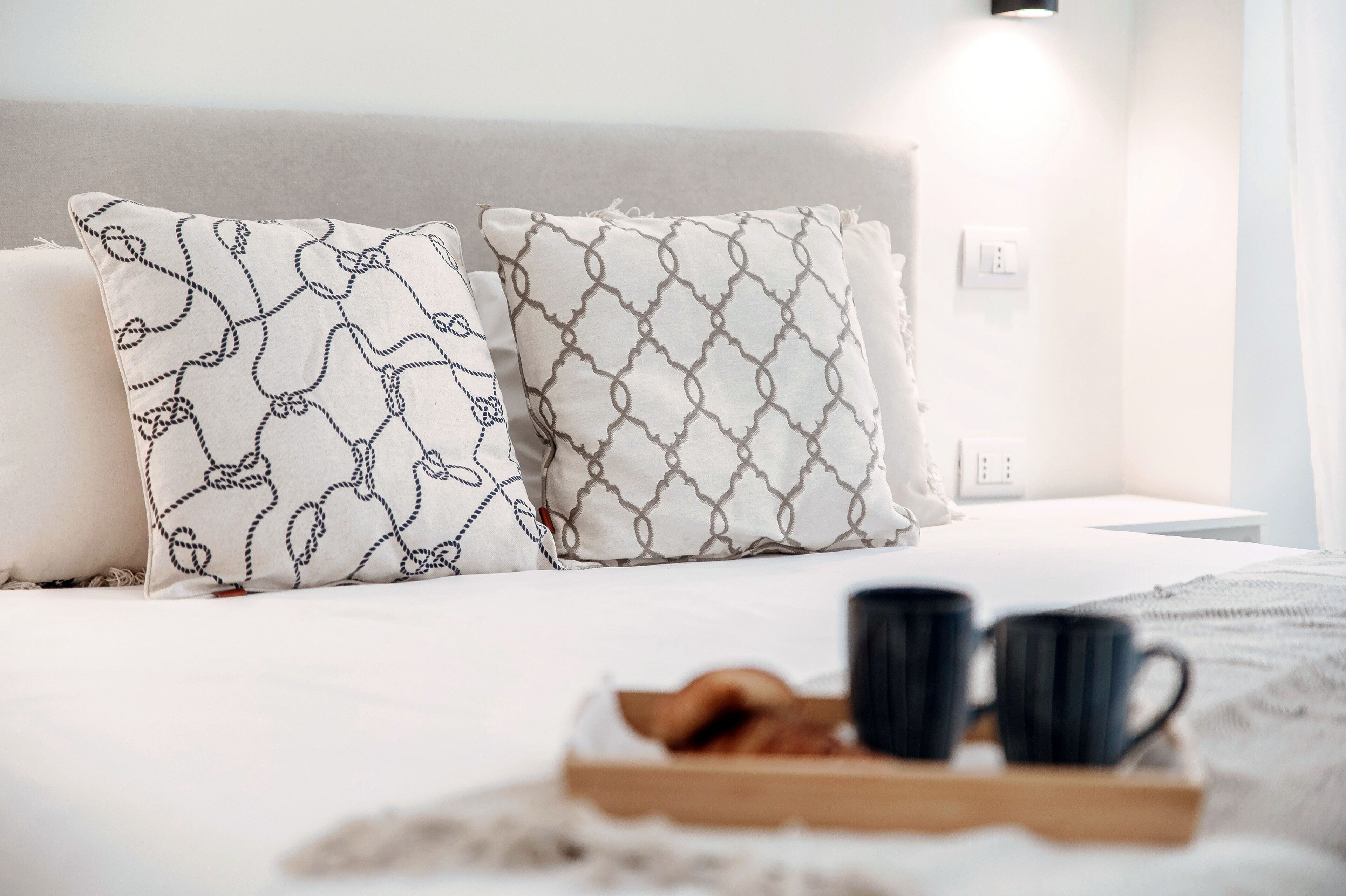 A neatly made bed with two patterned pillows, a tray with croissants and black mugs, and a wall-mounted light.