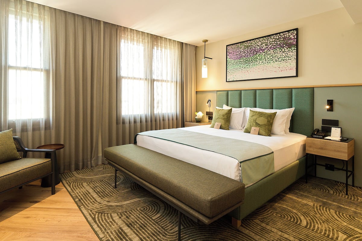 A hotel room with a large bed, a bench, a bedside table with a lamp, and a wall-mounted artwork.