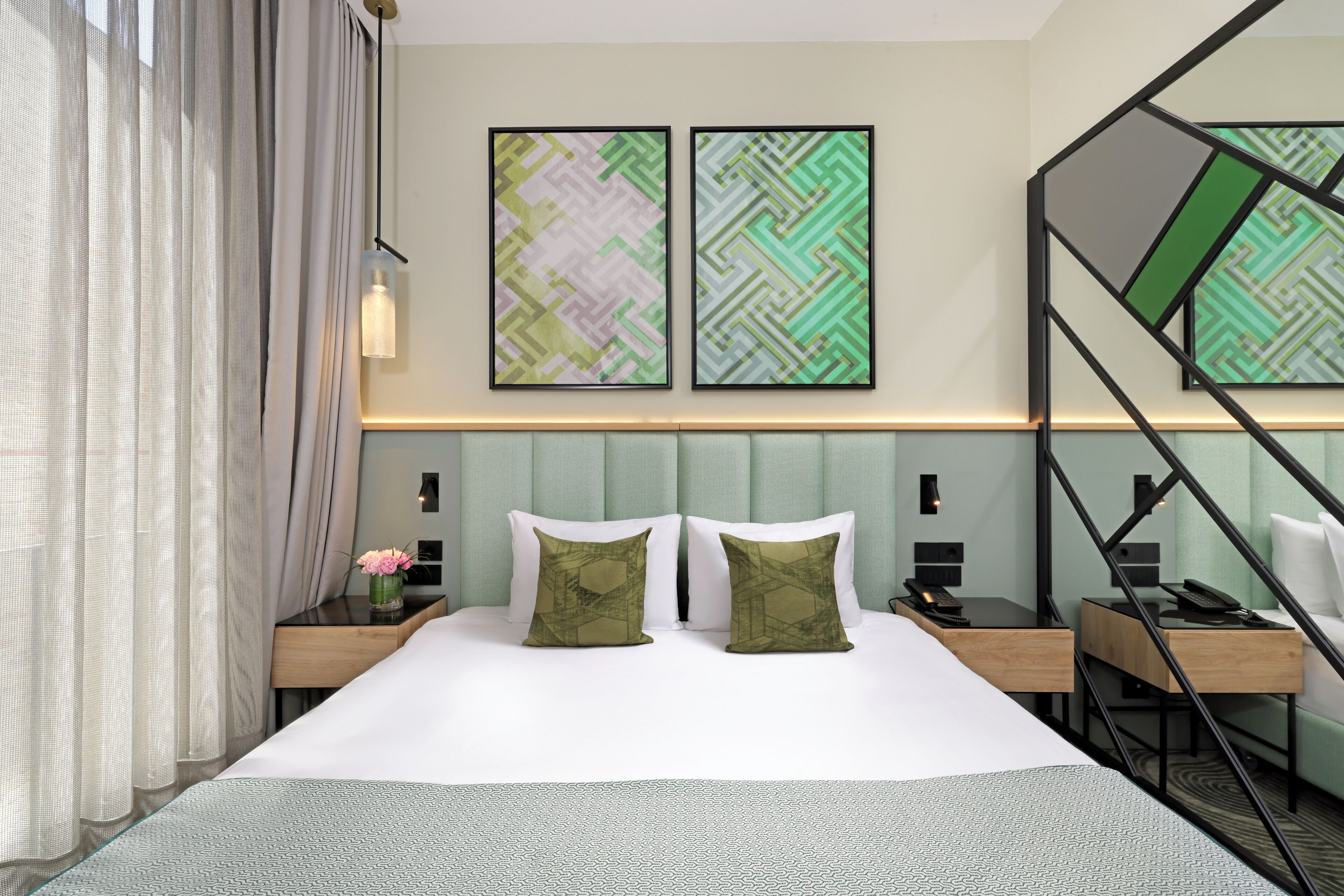 Ein Hotelzimmer mit einem Bett, zwei Nachttischen, einem Schreibtisch und einem Sessel. An der Wand über dem Bett hängen zwei gerahmte abstrakte Gemälde.