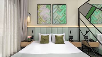 Ein Hotelzimmer mit einem Bett, zwei Nachttischen, einem Schreibtisch und einem Sessel. An der Wand über dem Bett hängen zwei gerahmte abstrakte Gemälde.