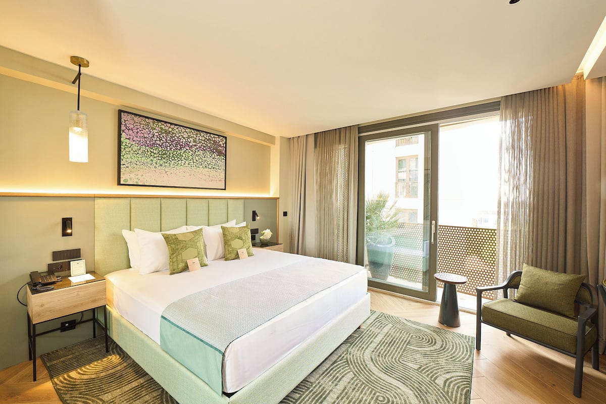 A hotel room with a large bed, a nightstand, a chair, a small table, and a view of the outside through a sliding glass door.