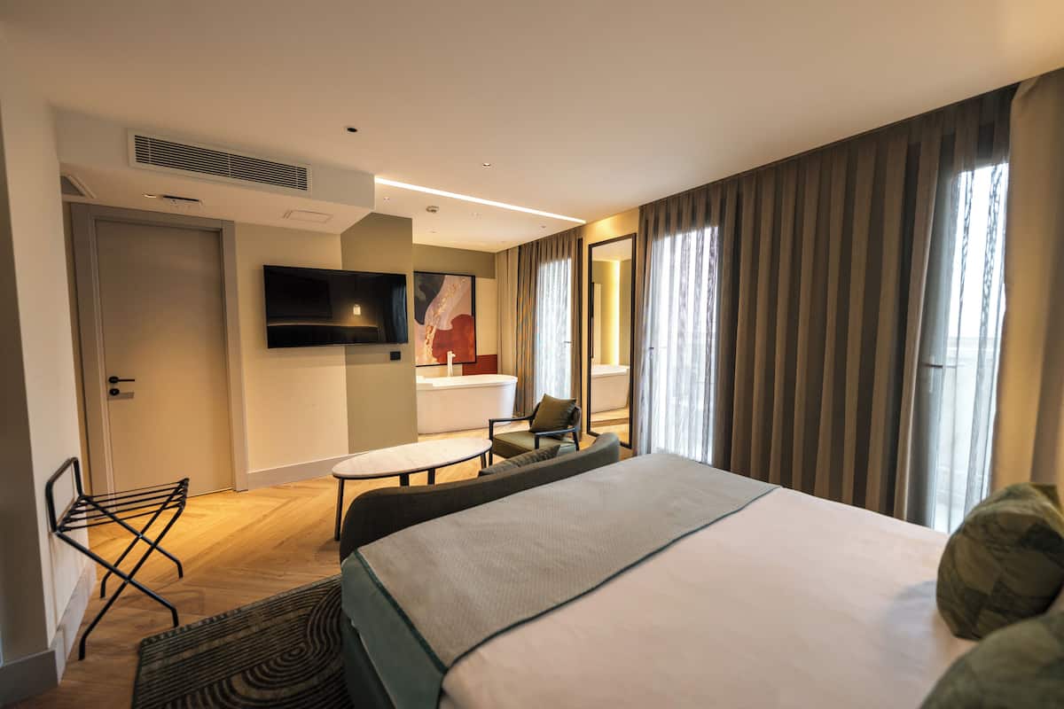 A hotel room with a bed, a seating area with a table and chairs, a television, and a large window with curtains.