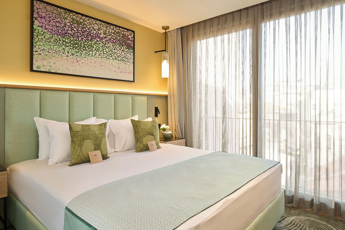A hotel room with a large bed, a headboard, a bedside table with a lamp, and a window with sheer curtains.