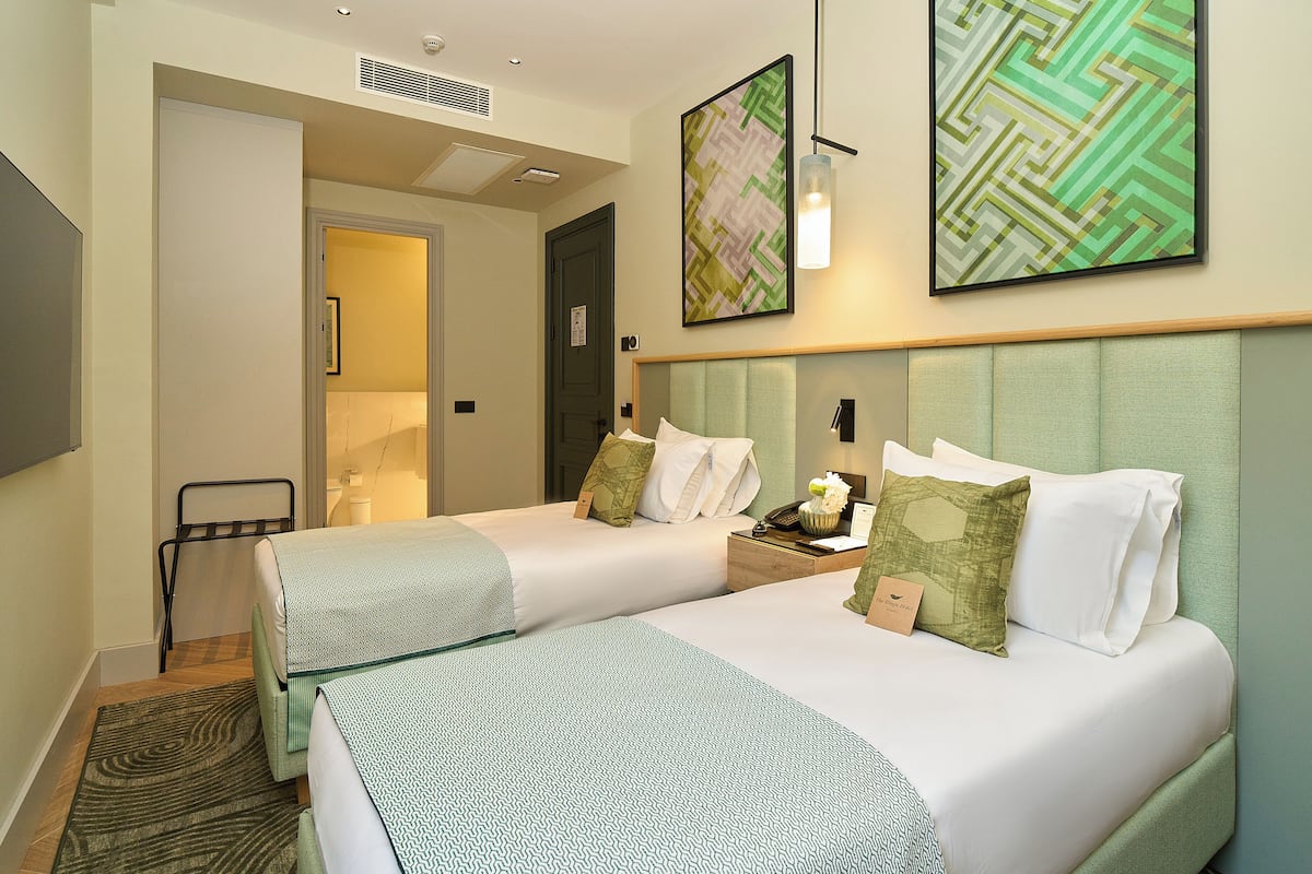 A hotel room with two beds, a nightstand, a bedside table, a wall-mounted artwork, and a ceiling vent.