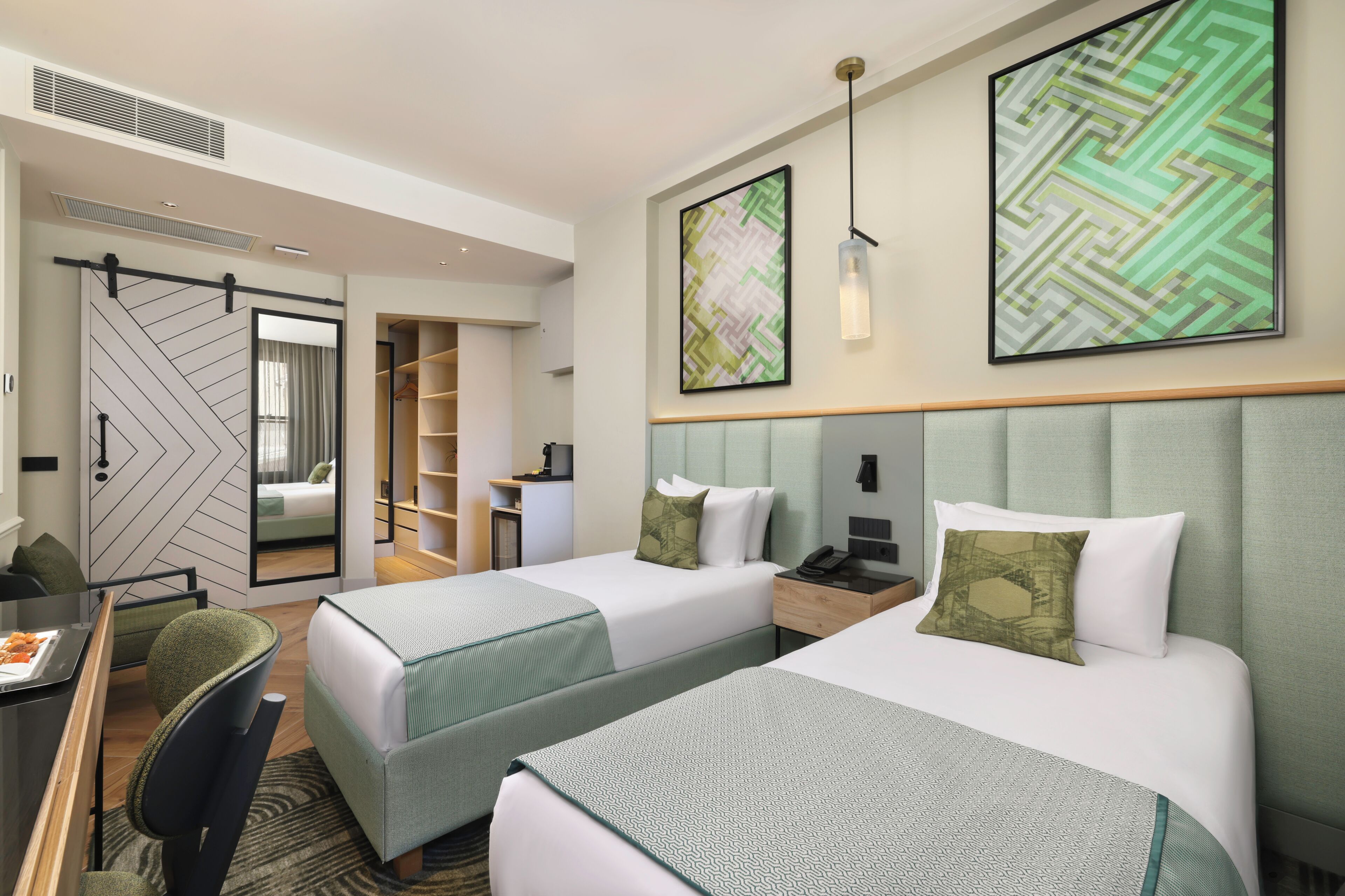A hotel room with two beds, a desk, a chair, a wardrobe, and artwork on the walls.