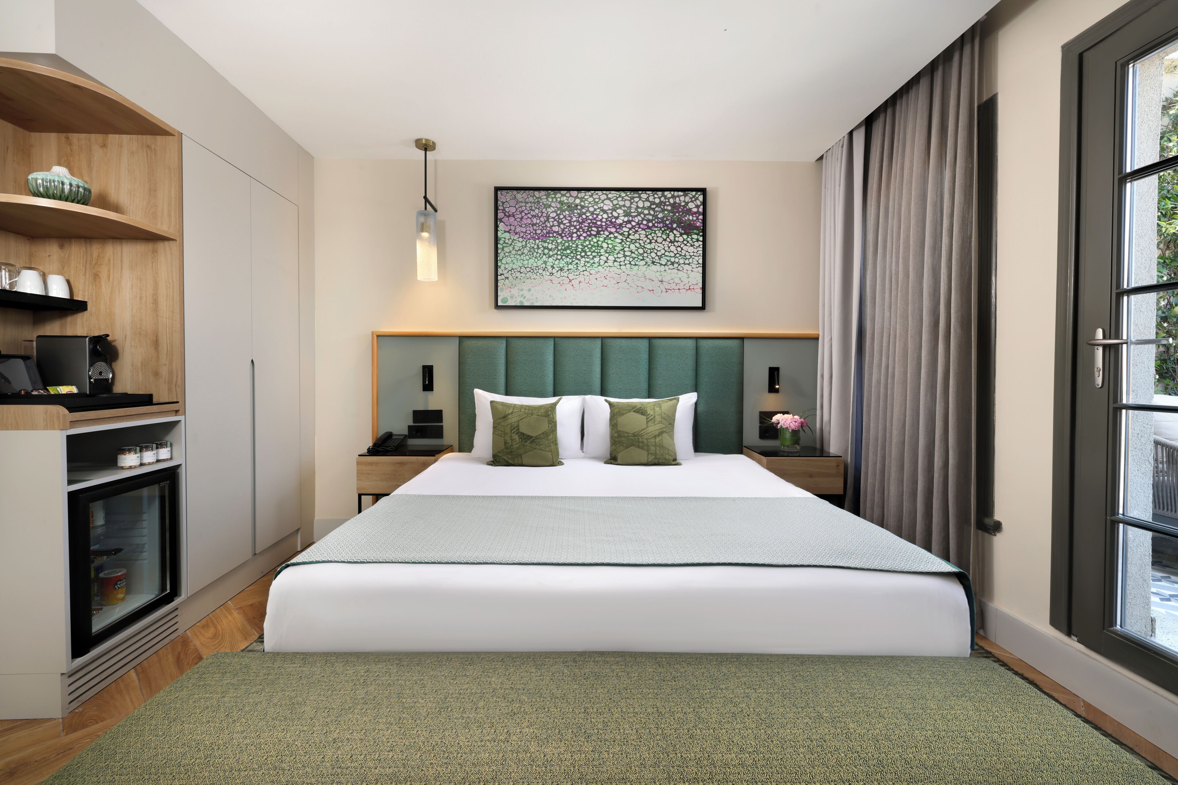 A hotel room with a bed, a desk, a refrigerator, a window with curtains, and a wall-mounted artwork.