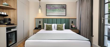 A hotel room with a bed, a desk, a refrigerator, a window with curtains, and a wall-mounted artwork.