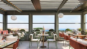 Un área de comedor al aire libre con mesas y sillas, amplias ventanas con vista a la ciudad y lámparas colgantes.