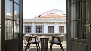 Un balcón abierto con dos sillas y una mesita, con vistas a un edificio de techos con tejas y paredes blancas.