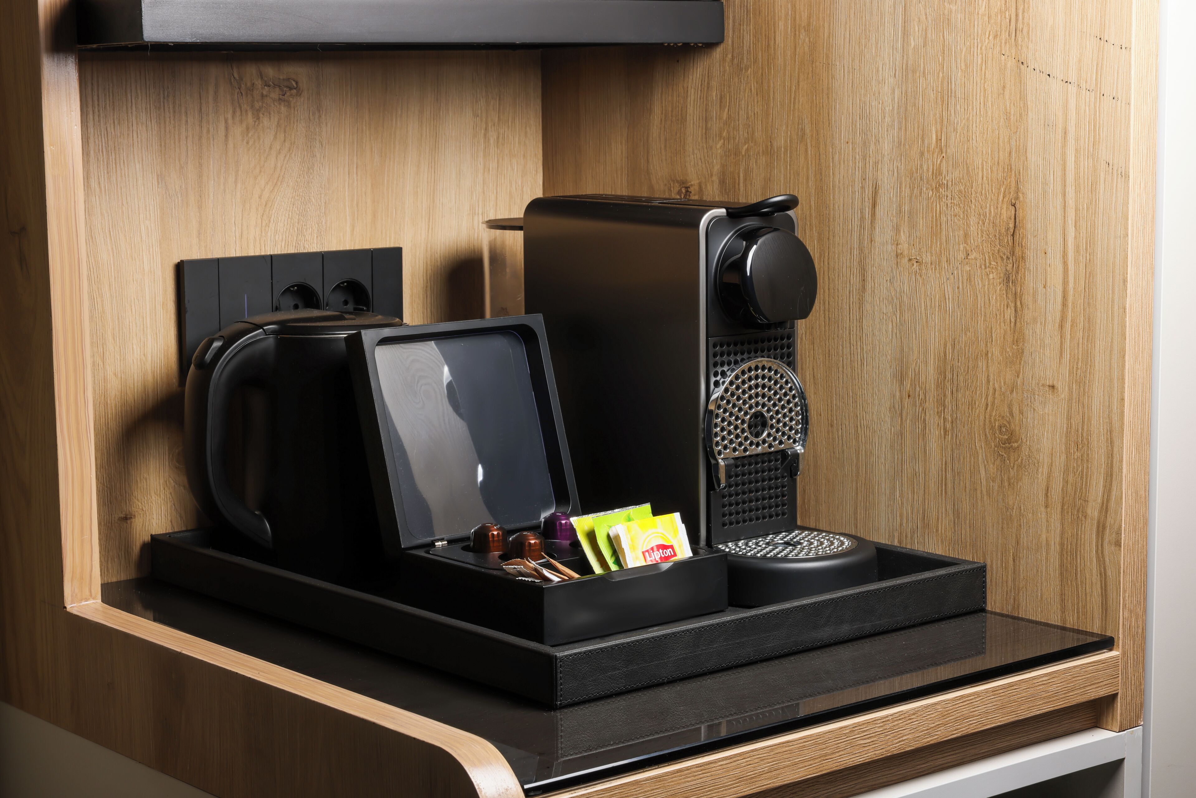 Une machine à café avec un bac de récupération, une bouilloire et un support pour capsules de café, le tout posé sur un comptoir en bois.
