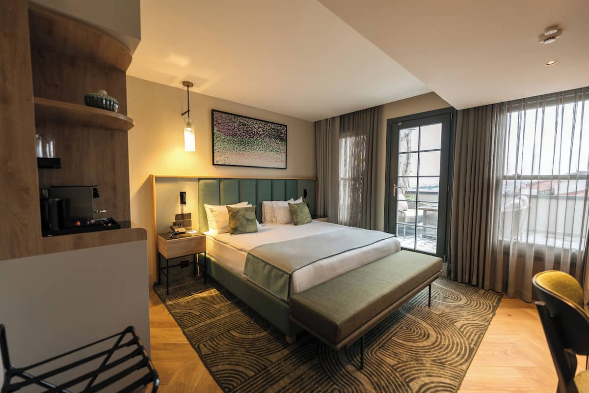 A modern hotel room with a large bed, a desk, a chair, and a view of the outdoors.