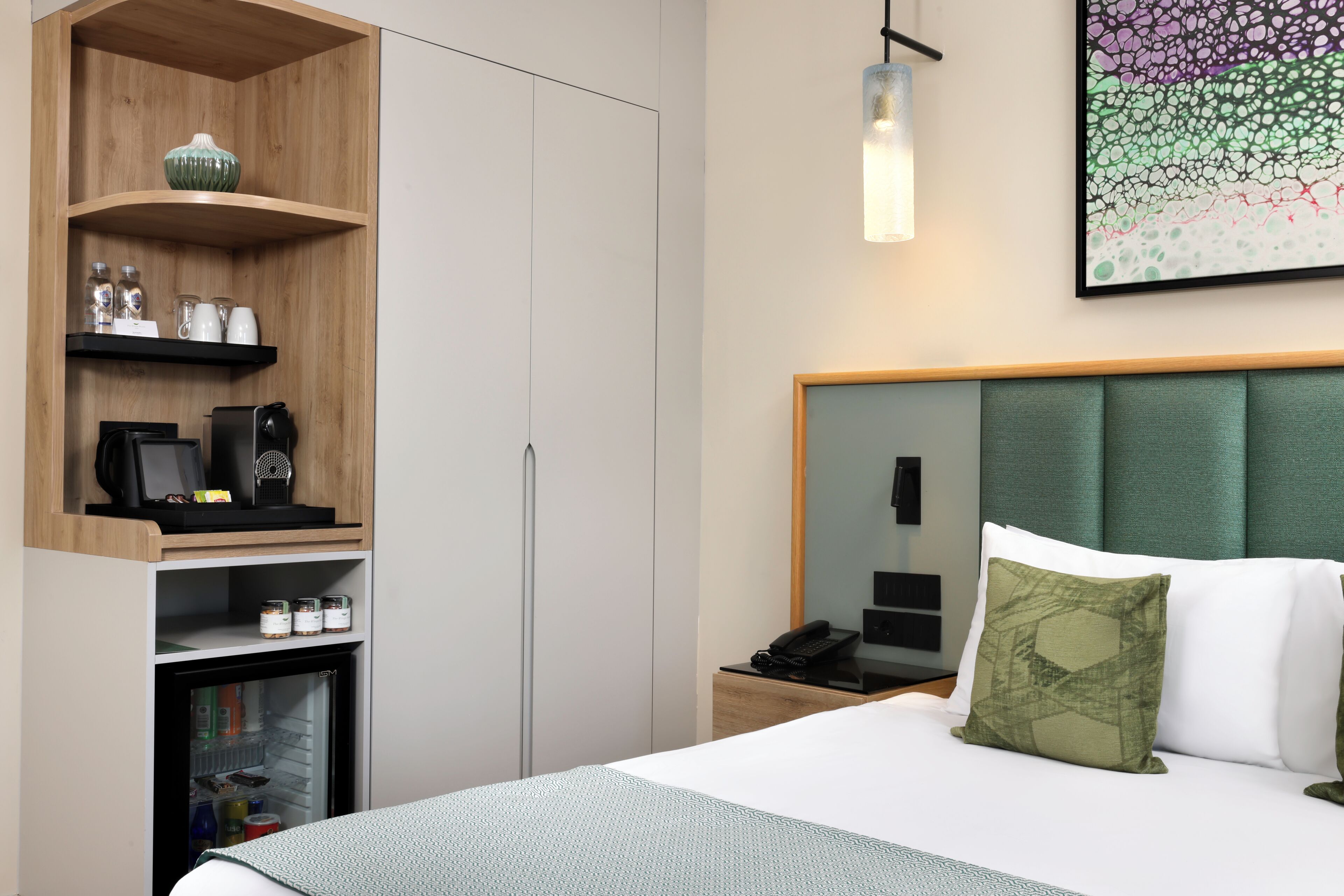 A hotel room with a bed, a nightstand, a coffee machine, a refrigerator, and a wall-mounted artwork.