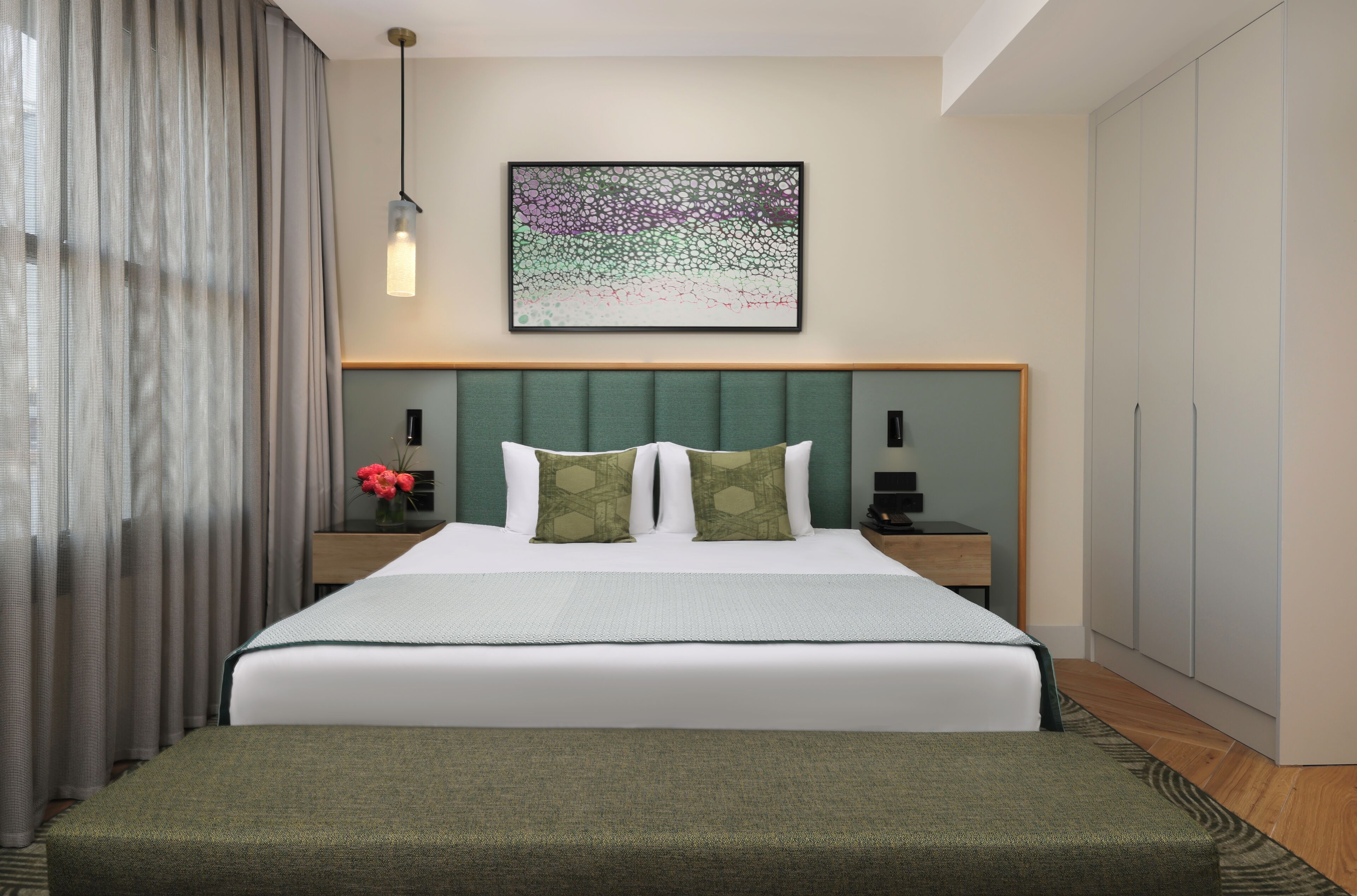 A modern hotel room with a large bed, a green headboard, a bedside table with a lamp, and a framed abstract artwork on the wall.