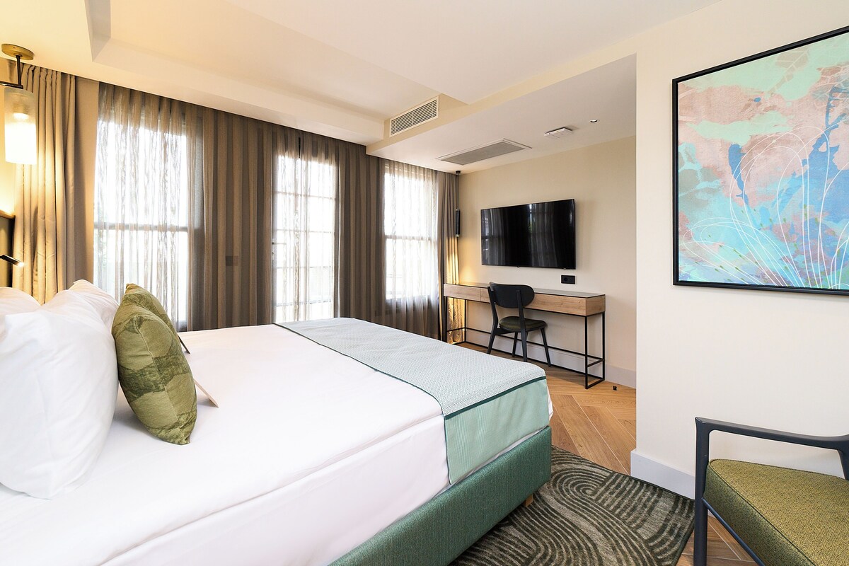 Habitación de hotel con cama, escritorio, silla, televisor y ventana grande con cortinas.