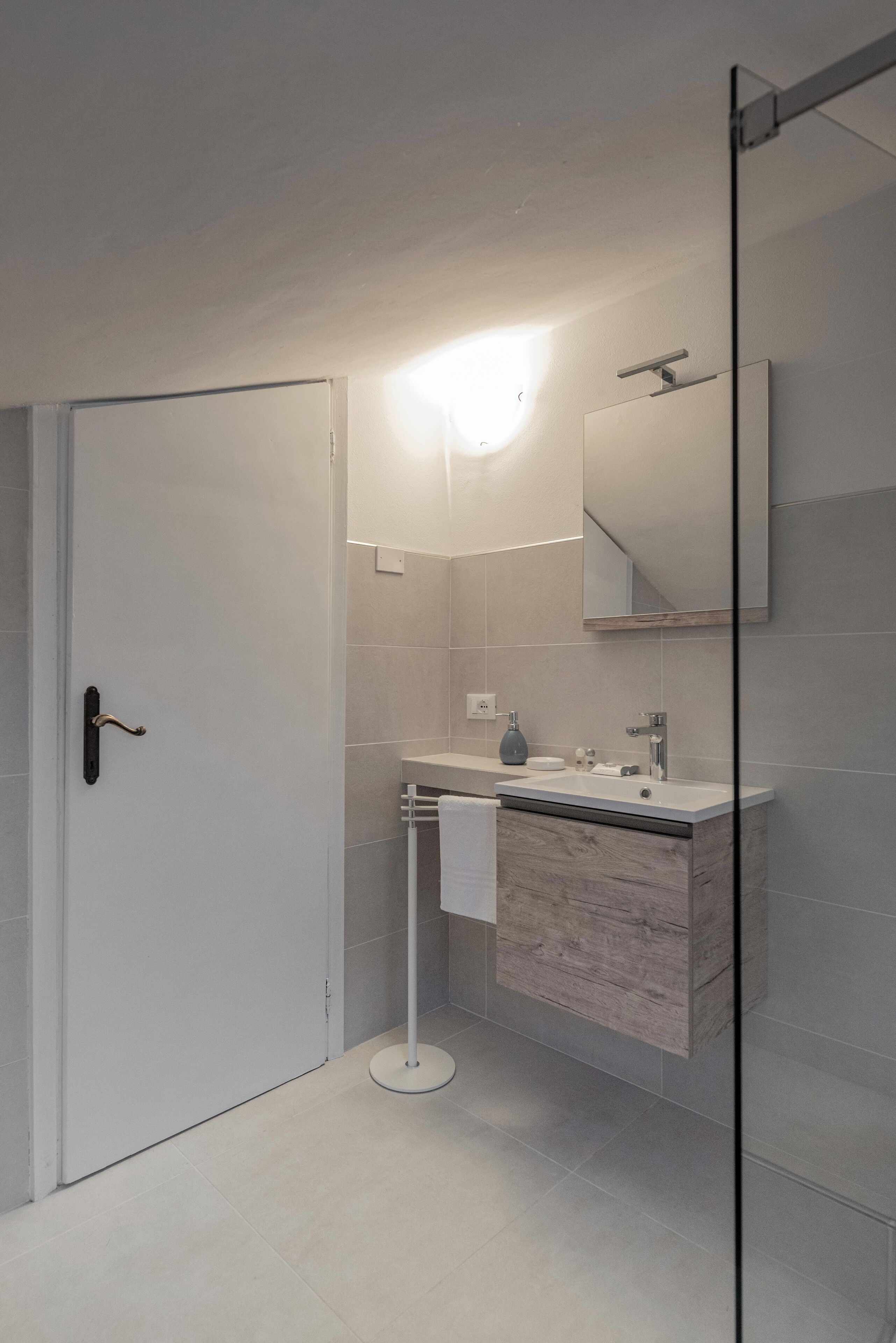 A modern bathroom with a white door, a glass shower enclosure, a white sink with a wooden vanity, and a mirror above the sink.