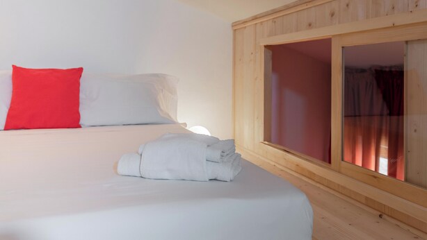 A bed with white bedding and a red pillow, a rolled-up towel, and a wooden-framed window with a view of a red curtain. - EnJoy Home - Attic with terrace in downtown/hospital area (Padova)