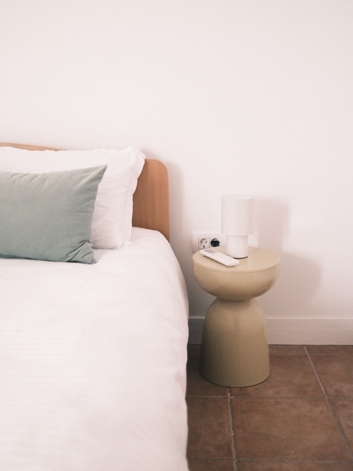 A bed with white bedding and a green pillow, a bedside table with a white lamp, and a wall socket.
