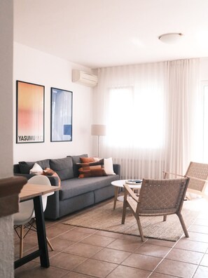 Ein modernes Wohnzimmer mit einer Couch, einem Couchtisch und zwei Sesseln. An der Wand hängen zwei gerahmte Bilder, eine Lampe und ein Fenster mit Vorhängen.