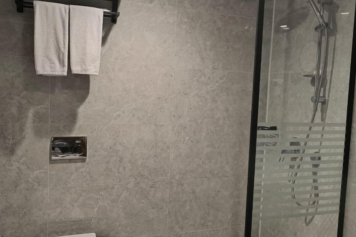 Un baño con cabina de ducha de vidrio, un perchero con dos toallas y un pequeño estante con un espejo.