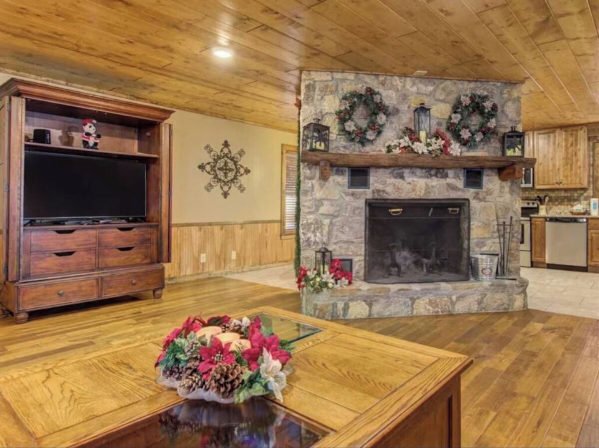 A cozy living room with a stone fireplace, wooden entertainment center, and a glass coffee table with a floral arrangement.