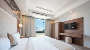 A hotel room with a large bed, a flat-screen TV mounted on a wooden panel, a balcony with curtains, and a view of the cityscape.