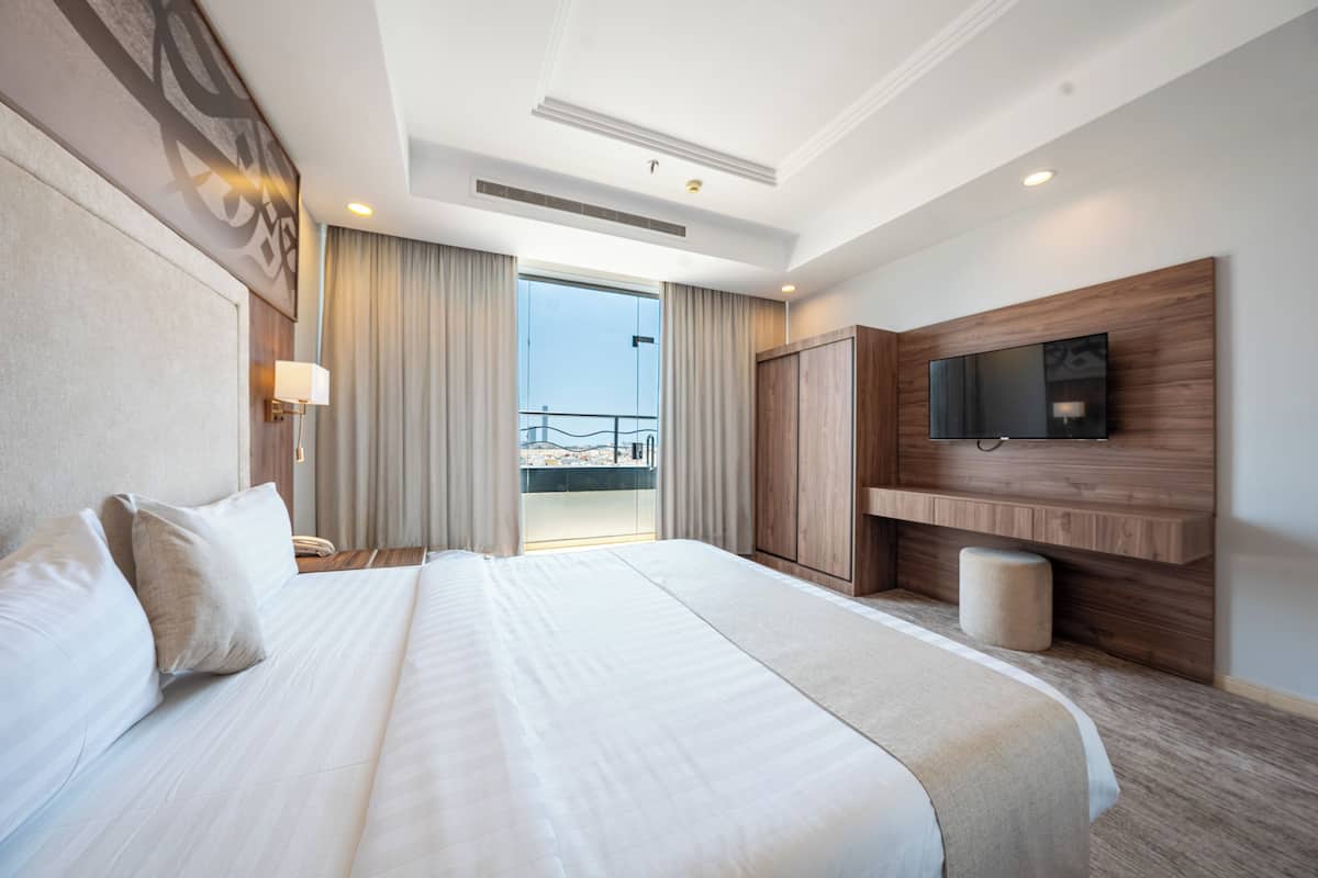 Une chambre d’hôtel dotée d’un grand lit, d’un téléviseur à écran plat fixé sur un panneau en bois, d’un balcon avec des rideaux et offrant une vue sur le paysage urbain.