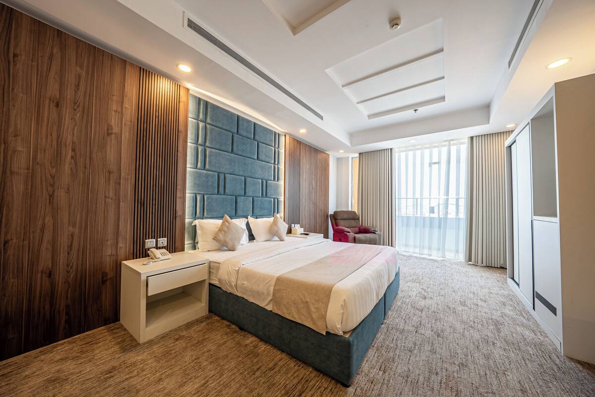 Une chambre d’hôtel moderne avec un grand lit, une table de chevet, une tête de lit intégrée et une grande fenêtre avec des rideaux.
