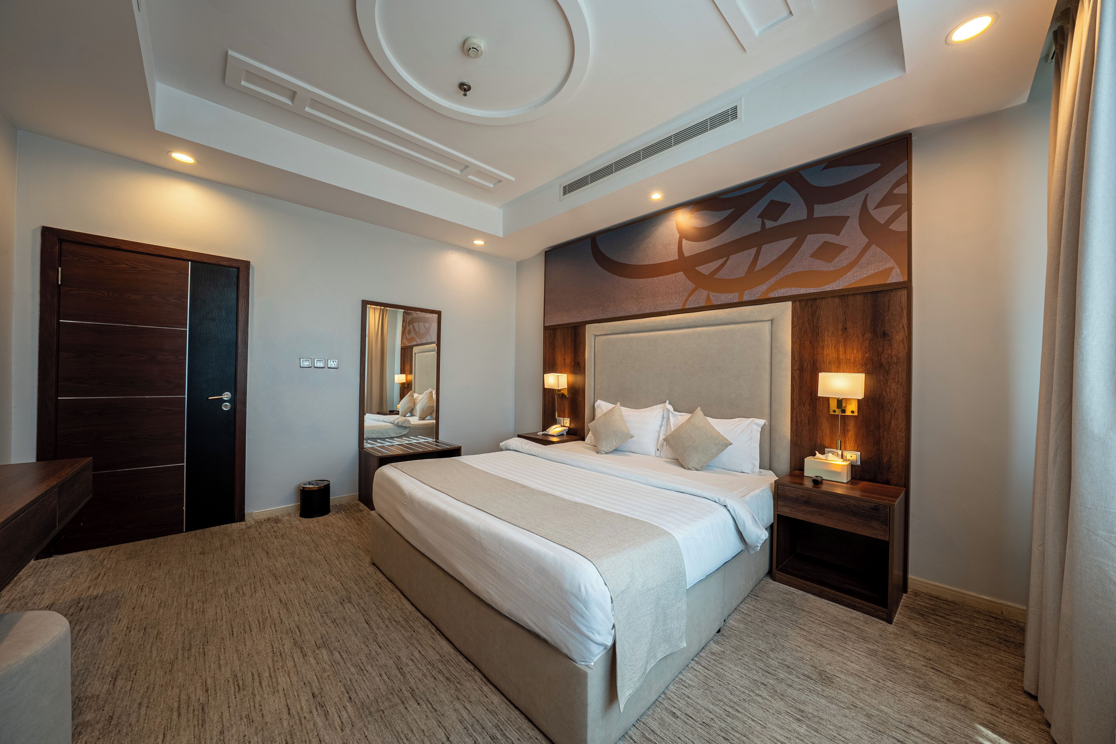 Une chambre d’hôtel avec un grand lit, des tables de chevet, un miroir et une tête de lit en bois.