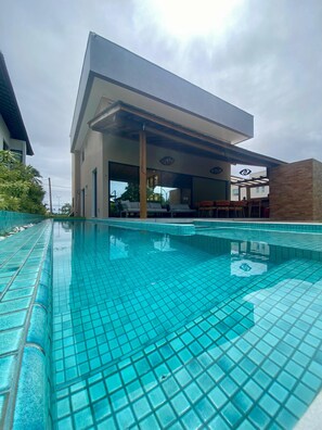 A swimming pool with a tiled deck, a covered seating area with chairs, and a building with a modern design in the background. - Casa 81 Luxo Pé na Areia Muro Alto byLP (Ipojuca)
