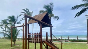 Estructura de juego de madera con tobogán y columpios, rodeada de césped y palmeras, con vistas a la playa.